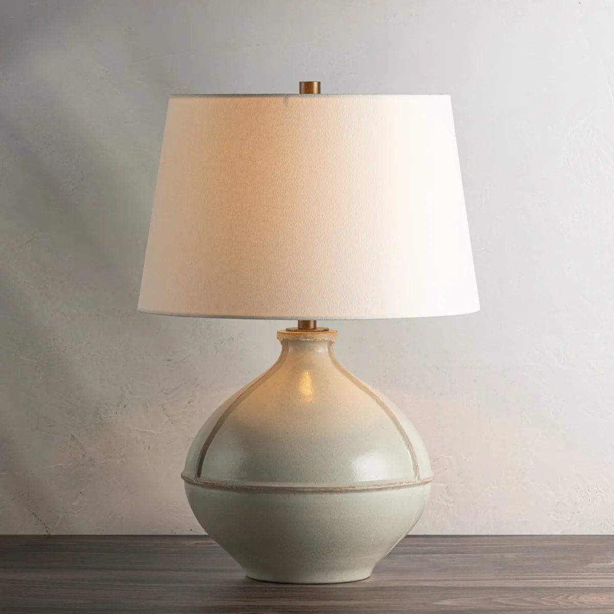 Inspired by rustic pottery, this table lamp's beautifully shaped base is crafted of pale sage ceramic giving it an earthy pale green tone. The off white linen shade pairs naturally and casts a soft glow. Raised seamlines and a reactive glaze that allows hints of the natural ceramic to peek through, give the fixture a hand-thrown, crafted feel. Amethyst Home provides interior design, new home construction design consulting, vintage area rugs, and lighting in the Kansas City metro area.